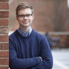 A white man with glasses wearing a blue sweater has his arms crossed. He is smiling and leaning on a brick wall.